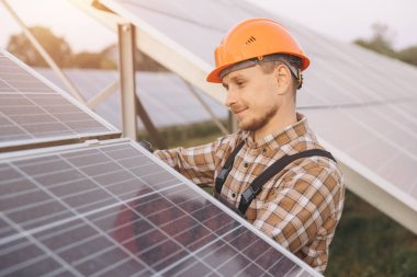 Erkek bir mühendis güvenlik kaskı takarak güneş panellerini inceliyor. Güneşin altında çalışıyor, verimli güneş enerjisi üretimi ve sürdürülebilirliğini sağlıyor..