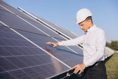 Engineer inspects solar panels under bright sun, highlighting focus on sustainable energy solutions. Wearing a hard hat signifies safety in renewable energy development. clipart