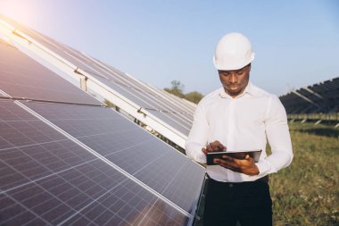 Sert şapkalı Afrikalı Amerikalı mühendis dışarıda dijital tablet kullanarak güneş panellerini inceliyor. Sahne, güneş çiftliği ortamında yenilenebilir enerji, teknoloji ve sürdürülebilir kaynakları vurguluyor.