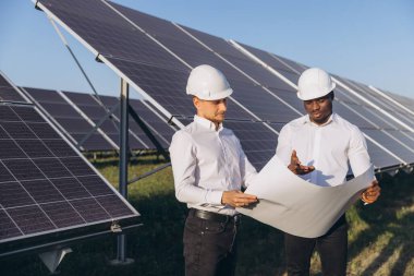 Sert şapkalar takan iki mühendis, yenilenebilir enerji ve işbirliğini vurgulayarak bir güneş çiftliğinde mimari planları tartışıyor. Güneş ışığı, güneş teknolojisinin verimliliğini ve potansiyelini vurguluyor.