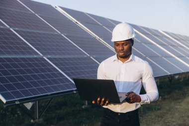 Sert şapka takan Afrikalı Amerikalı mühendis dizüstü bilgisayar kullanarak güneş panellerini inceliyor. Bu resim yenilenebilir enerji teknolojisini ve çevresel yenilikleri vurgular.