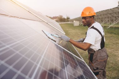 Koruyucu kıyafet giyen Afrikalı Amerikalı erkek teknisyen güneşli bir günde büyük güneş panellerini temizliyor ve sürdürülebilir enerji çözümleri için maksimum enerji verimliliği ve performans sağlıyor..