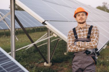 A confident worker in a checked shirt and hard hat stands with folded arms by solar panels. This image captures the essence of renewable energy and professional expertise in solar power technology. clipart