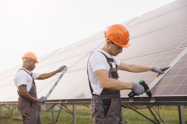 Güvenlik donanımlı iki işçi yenilenebilir enerji ve takım çalışmasını vurgulayarak güneş panelleri kuruyor. Görüntü sürdürülebilirlik, yenilik ve çevresel sorumluluğu güneşli ve açık ortamda taşır..