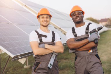 Sert şapkalı iki mühendis, bir kurulum projesi sırasında, takım çalışması, yenilenebilir enerji ve sürdürülebilir çözümleri temsil eden güneş panelleri önünde durarak güvenle gülümsüyor..