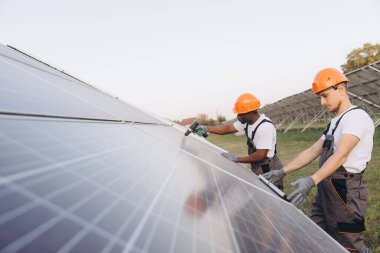 İki güvenlik ekipmanı mühendisi dışarıda güneş panelleri kurmaya çalışıyor. Açık mavi gökyüzü altında, yenilenebilir enerji ve sürdürülebilir uygulamalara olan bağlılıklarını ve takım çalışmalarını gösteriyorlar..
