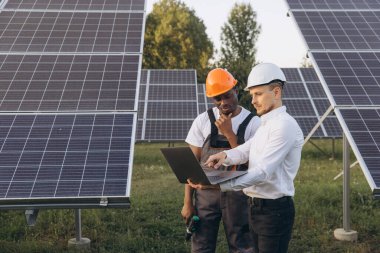 Two engineers wearing hard hats work together with a laptop in a solar panel field. They focus on efficient energy solutions and renewable power, showcasing teamwork and sustainable technologies. clipart