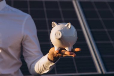 Bir işadamı arka planında güneş panelleri olan bir kumbara tutuyor. Yatırımı, tasarrufu ve yenilenebilir enerjiyi simgeliyor. Görüntü, mali planlama ve sürdürülebilir büyüme kavramını aktarıyor.