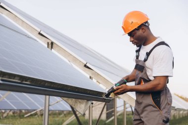 Güvenlik ekipmanlı Afrikalı Amerikalı teknisyen güneş panellerini açık havada, güneşli bir ortamda kurmaya çalışıyor. Yenilenebilir enerji, sürdürülebilir teknoloji ve çalışkan profesyonellik.