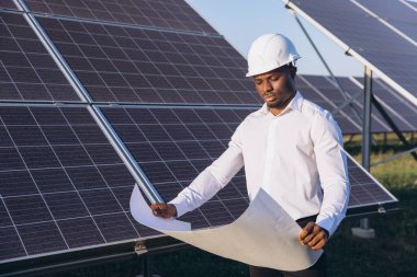 Sert şapka takan Afrikalı Amerikalı mühendis, yenilenebilir enerji ve sürdürülebilir teknolojiyi vurgulayarak, büyük güneş panellerinin yanında dururken bir planı inceliyor..