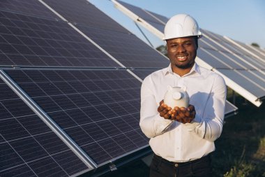 Sert şapkalı gülümseyen bir Afrikalı Amerikalı mühendis, yenilenebilir enerji tasarrufu ve yatırımı simgeleyen güneş panelleri önünde kumbara tutuyor. Sahne sürdürülebilirlik ve mali büyüme gösteriyor.