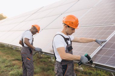 Koruyucu teçhizat kullanan iki mühendis yenilenebilir enerji alanına güneş panelleri yerleştirmeye çalışıyor. İşbirliği gösteriyorlar ve sürdürülebilir teknolojiye odaklanıyorlar. Ayar güneşli bir açık hava yeri.