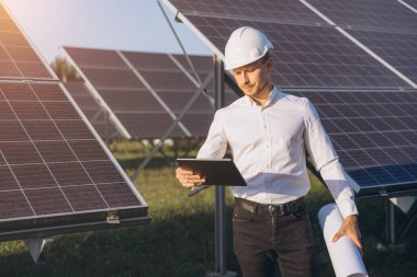 Sert şapka takan erkek bir mühendis güneş panellerini bir güneş çiftliğinde inceliyor. Yenilenebilir enerji teknolojisi ve sürdürülebilir çözümleri vurgulayan bir tablet ve plan elinde..