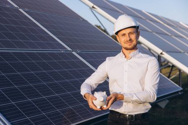 A confident engineer wearing a hard hat smiles while holding a piggy bank in front of solar panels, symbolizing renewable energy savings and sustainable technology innovation. clipart