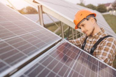 Sert şapka takan bir teknisyen yenilenebilir enerji çiftliğindeki güneş panellerini inceliyor. Batan güneş, sürdürülebilir enerji çözümleri ve yenilikçiliğe odaklanarak sıcak bir ışık saçıyor..