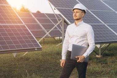 Sağlam bir şapka takan ve dizüstü bilgisayar tutan kendinden emin bir mühendis güneş panelleri arasında duruyor, yenilenebilir enerji yeniliği ve güneş enerjisi çiftliği ortamında sürdürülebilirliği temsil ediyor..