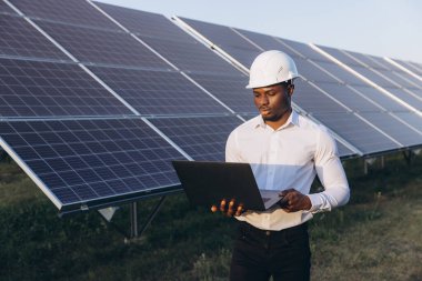 Beyaz kasklı Afrikalı Amerikalı mühendis yenilenebilir enerji ve teknolojiyi vurgulayarak güneş panelleri önünde dizüstü bilgisayarda çalışıyor. Sahne bir yenilik ve sürdürülebilirlik hissi veriyor.
