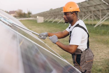 African American Male Engineer wearing safety gear performs maintenance on solar panels at a solar farm. Image captures renewable energy, sustainable technology, and professional work in progress. clipart