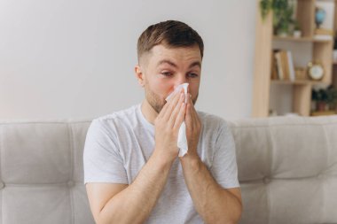 Mutsuz orta yaşlı adam ateş ve gripten muzdarip, burnu peçeteye sümküren, oturma odasında iç tarafta oturan. Covid-19 tecrit, hastalığın tedavisi, soğuk ve sulu.