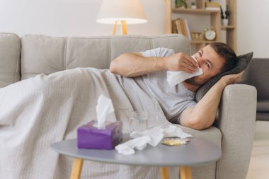 Evdeki kanepede sıcak bir battaniyenin altında yatıp kağıt mendille hapşıran hasta, beyaz bir adam. Soğuk algınlığı, grip, mevsimsel kış enfeksiyonları