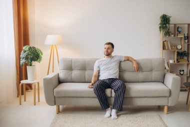 Evdeki oturma odasındaki kanepede oturan pijamalı neşeli, sakallı adam.