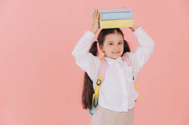 Sırt çantalı, pembe arka planda kitaplarını tutan, üniformalı, sevimli, güler yüzlü bir kız öğrenci.