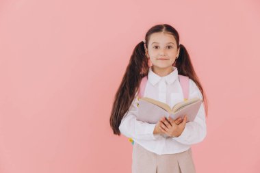 Sırt çantasıyla pembe arka planda kitap okuyan, okul üniformalı tatlı, güler yüzlü küçük kız.