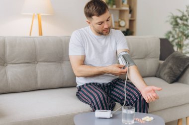 Evdeki kanepede oturan genç beyaz adam, kan basıncını ölçüyor. Modern tonometre kullanan sakallı adam stres ve baş ağrısından kaynaklanan hipertansiyon yaşıyor.