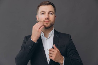 Saçını ve sakalını tarayıp kameraya bakan gri takım elbiseli sakallı iş adamının portresi. Gri stüdyo arkaplanı üzerinde kopya alanı olan bir portre.