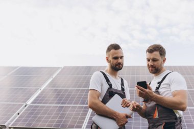 Güneş paneli inşaatının verimliliğini kontrol etmek için dijital akıllı telefon okuma bilgilerini kullanan bir enerji uzmanı. Yeşil enerji işleri. Teknoloji. İki güneş enerjisi santrali işçisi veri kaydediyor