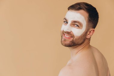 Tıraşsız, gülümseyen bir adamın güzel portresi bej renkli stüdyo arka planına yüz bakımı maskesi takıyor. Dermatoloji, lüks spa prosedürleri. Erkek güzellik anlayışı, kozmetik ürünler..