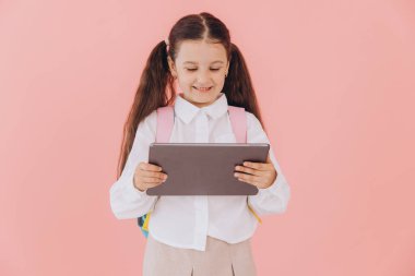 Sırt çantasında pembe arka planda tablet taşıyan, okul üniformalı sevimli, küçük bir kız.