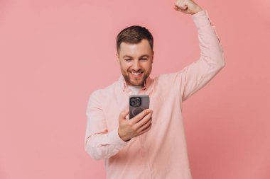 Bir kazananımız var! Pembe tişörtlü mutlu bir genç zafer ifadesiyle telefon ekranına bakıyor, pembe eğimli arka planda izole edilmiş.