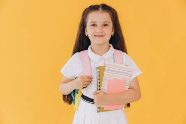 Sırt çantalı, sarı arka planda defterleri ve kitapları olan beyaz okul üniformalı küçük tatlı kız.