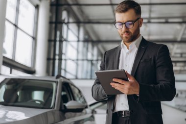 Sakallı, gözlüklü, takım elbiseli, araba tamirhanesinde duran ve elinde tablet tutan dost canlısı araba satıcısı.