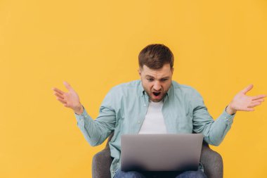 Tıraşsız, naneli gömlekli bir adam sandalyede oturur dizüstü bilgisayarda çalışır ve ellerini sarı arka plan stüdyosuna yayar..