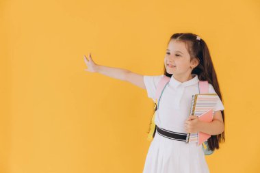 Beyaz okul üniformalı, elinde not defterleri olan ve sarı arka planda eli olan küçük tatlı bir kız.