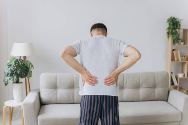 Olgun bir adamın beyaz pijama giyip, sabah evde dikilip, sırtına dokunarak kas gerginliğinden muzdarip olduğu arka plan görüntüsü.