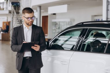 Yakışıklı, sakallı araba satıcısı takım elbiseli bir klasör tutuyor ve arabanın yanında dururken gülümsüyor.