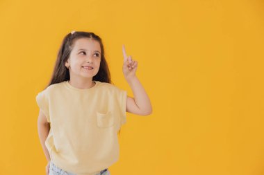 Küçük şirin bir kızın portresi bir şey gösteriyor ve düşünüyor, sarı arka planda fotokopi alanı ile izole edilmiş.