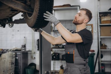 Yoğunlaştırılmış araba servisi çalışanının otomobil kontrol ekipmanlarına bakın. Üniformalı ve eldivenli erkek tamirci arabanın altında duruyor ve kırılma noktası arıyor.