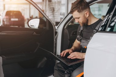 Otomotiv teknisyeni bilgisayarda tanılama yazılımı çalıştırıyor. Tamirci, oto servis garajında aracın tanılama testini yaparken arabada oturup dizüstü bilgisayarla çalışıyor..