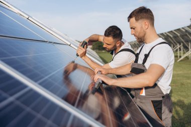 Gri üniforma giyen iki yetenekli işçi veya zanaatkar, teknisyenler temiz enerji ve elektrik tedariği için güneş panelleri kuruyorlar.