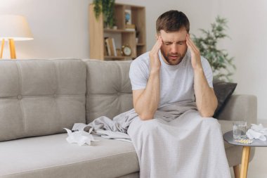 Ateşli ve baş ağrısı çeken hasta bir adam kanepede otururken başını tutuyor. Tedavi ve hastalık kavramı