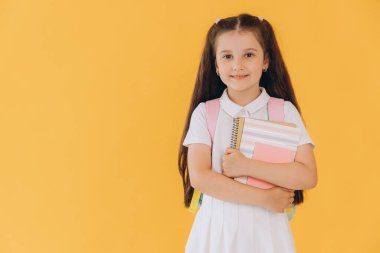 Sırt çantalı, sarı arka planda defterleri ve kitapları olan beyaz okul üniformalı küçük tatlı kız.