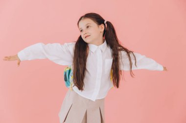 Beyaz tişörtlü, bej etekli, sırt çantalı pembe stüdyo arka planlı neşeli ilkokul öğrencisinin stüdyo portresi.