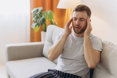 Evde baş ağrısı çeken genç bir adam, şakaklarına dokunuyor, fotokopi odası, bulanık arka plan. Migren, baş ağrısı, stres, gerilim sorunu, akşamdan kalma konsepti
