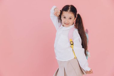 Sırt çantasıyla pembe arka planda yüzen, okul üniformalı tatlı, güler yüzlü küçük kız.