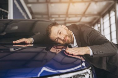 Pozitif, genç, sakallı, beyaz bir adam yeni arabasını bayilikte kucaklıyor. Mutlu adam sonunda resmi takım elbise içinde uzun zamandır beklenen arabaya kavuştu.