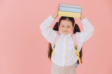 İlkokul öğrencisi elinde kitaplarla kameraya gülümsüyor. Sırt çantası ve pembe arka planda beyaz gömlek giyiyor.